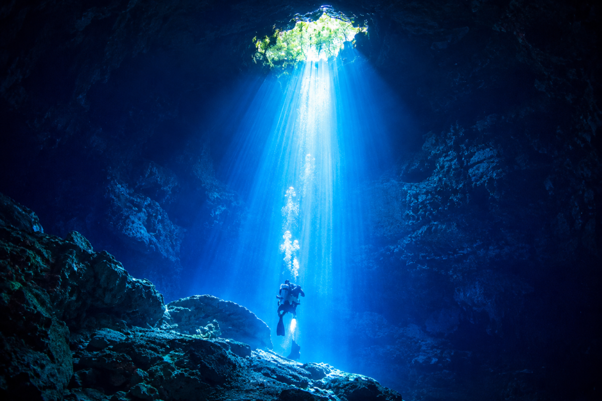Diver ascending beneath a dramatic shaft of sunlight inside The Pit Cenote near Tulum, Mexico.