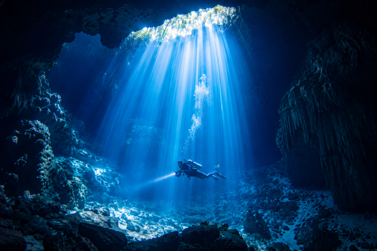Scuba diver swimming beneath dramatic light rays inside The Pit Cenote near Tulum Mexico.