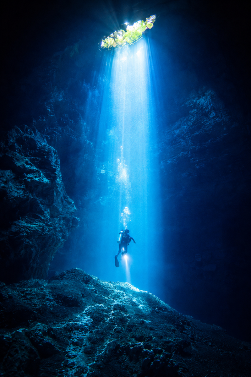 Scuba diver exploring The Pit Cenote beneath dramatic light beams and ancient stalactites near Tulum Mexico.